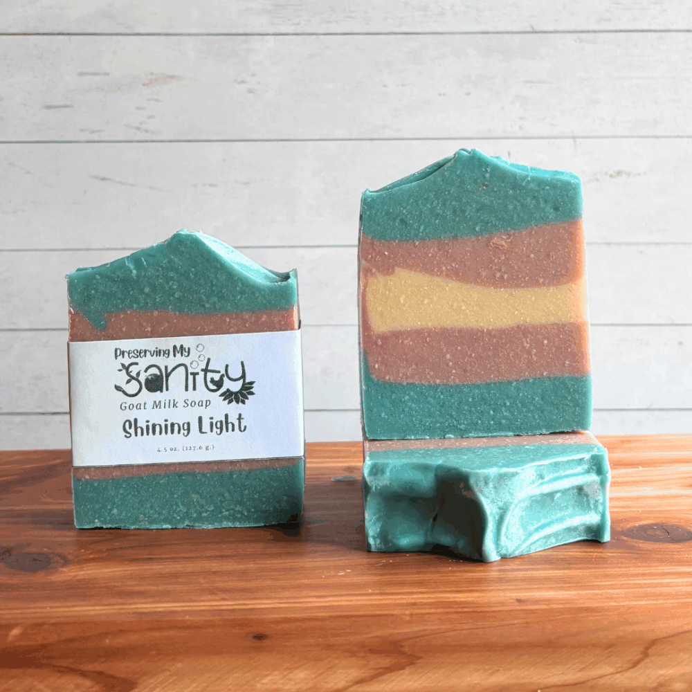 Three colorful bars of Swirl Power goat milk soap, featuring the blue, white, and pink colors of the Transgender flag in a striped pattern perfect for celebrating Pride Month. The left bar is labeled, while the other two are unlabeled to show top and face design. All are sitting on a cedar board with a white paneled background.