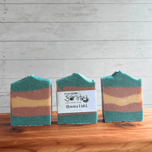 Three colorful bars of Shining Light goat milk soap, featuring the blue, white, and pink colors of the Transgender flag in a striped pattern perfect for celebrating Pride Month. The middle bar has a label, while the other two are unlabeled to show soap design. All are sitting on a cedar board with a white paneled background.