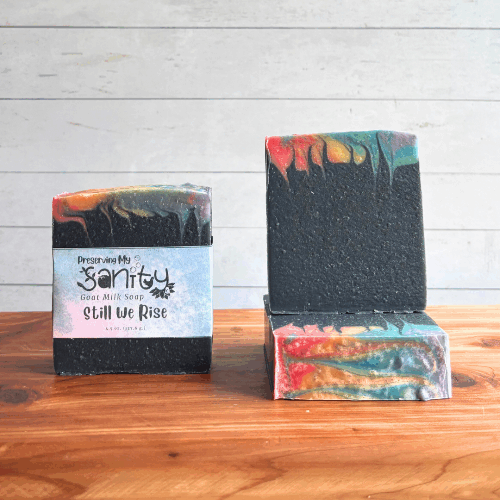 Three colorful rainbow bars of Swirl Power goat milk soap, featuring all seven colors of the Pride flag at the very top with the majority of the soap being black. The left bar is labeled, while the other two are unlabeled to show top and face design. All are sitting on a cedar board with a white paneled background.
