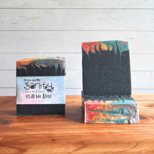 Three colorful rainbow bars of Swirl Power goat milk soap, featuring all seven colors of the Pride flag at the very top with the majority of the soap being black. The left bar is labeled, while the other two are unlabeled to show top and face design. All are sitting on a cedar board with a white paneled background.