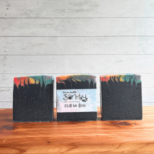 Three colorful rainbow bars of Still We Rise goat milk soap, featuring all seven colors of the Pride flag at the very top with the majority of the soap being black. The middle bar has a label, while the other two are unlabeled to show soap design. All are sitting on a cedar board with a white paneled background.