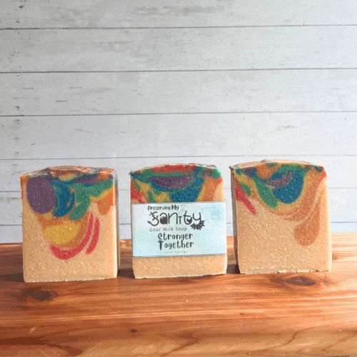 Three colorful rainbow bars of Stronger Together goat milk soap, featuring all seven colors of the Pride flag in a swirl pattern with a white base - perfect for celebrating Pride Month. The middle bar has a label, while the other two are unlabeled to show soap design. All are sitting on a cedar board with a white paneled background.