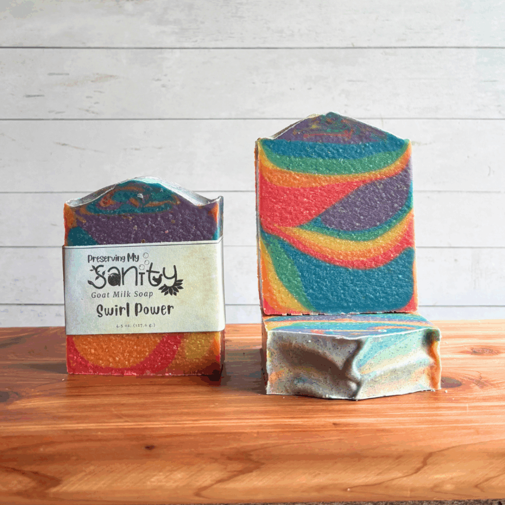 Three colorful rainbow bars of Swirl Power goat milk soap, featuring all seven colors of the Pride flag in a funky swirl pattern perfect for celebrating Pride Month. The left bar is labeled, while the other two are unlabeled to show top and face design. All are sitting on a cedar board with a white paneled background.