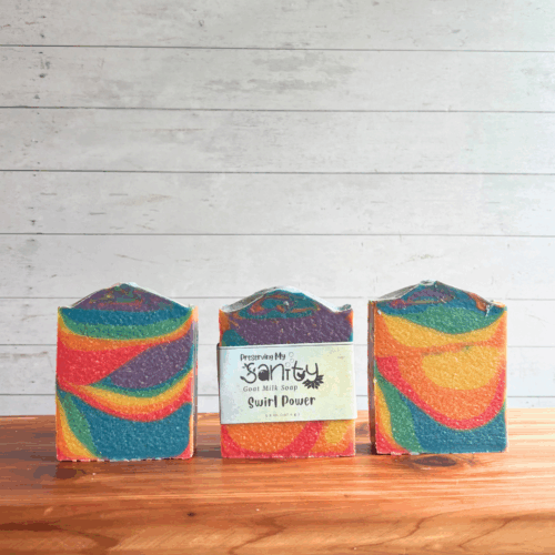 Three colorful rainbow bars of Swirl Power goat milk soap, featuring all seven colors of the Pride flag in a funky swirl pattern perfect for celebrating Pride Month. The middle bar has a label, while the other two are unlabeled to show soap design. All are sitting on a cedar board with a white paneled background.