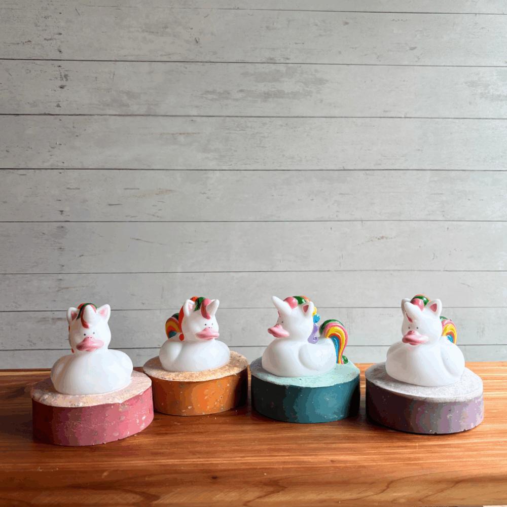Display photo showing four bars of Unicorn Poop soap with a toy unicorn on top, bars available in four colors - pink, orange, blue, and purple. Sitting on a cedar board with a white panel background.