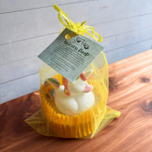 Display photo showing a fun bar of Unicorn Poop soap with a toy unicorn on top, packaged in a large cupcake liner inside a yellow organza bag and tied with a label. Sitting on a cedar board with a white panel background.