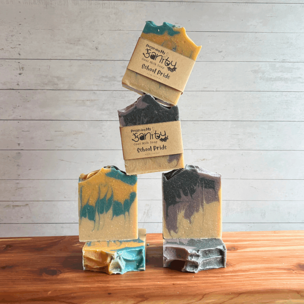 Fun display photo inspired by a cheerleader pyramid, showing six bars of School Pride goat milk soap. Three bars are black and purple, the other three are blue and gold. The top two that are balancing on the others are labeled, the others are unlabeled to show soap design and tops. All are sitting on a cedar board with a white panel background.