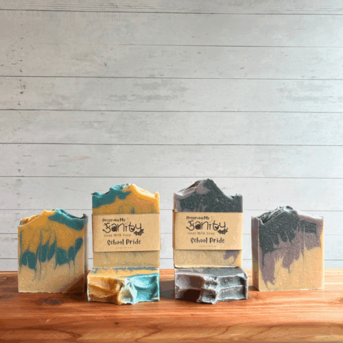 Display photo showing six bars of School Pride soap - three are blue and gold, three are black and purple. Two bars are labeled, and the other four are unlabeled to show soap design and tops. All are sitting on a cedar board with a white panel background.