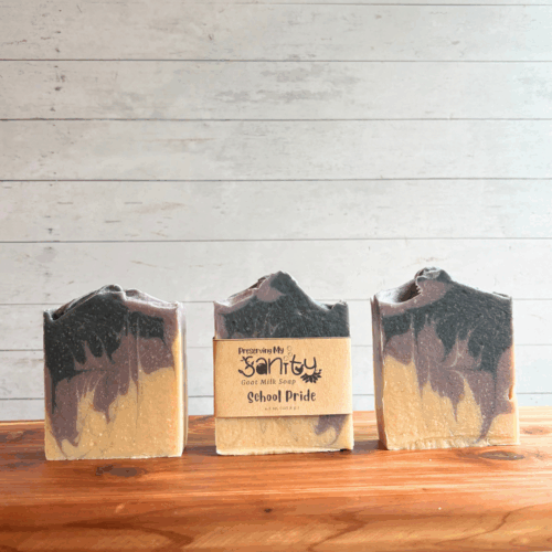 Display photo showing three bars of purple and black School Pride soap. The middle bar has a label, the other two bars are unlabeled to show soap design. All are sitting on a cedar board with a white panel background.