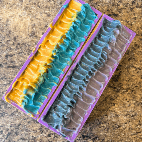 Process photo showing two loaves of freshly poured School Pride soap. One loaf is purple and black on top, the other is blue and gold.