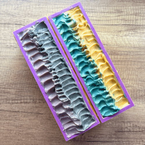 Process photo showing two loaves of School Pride soap before getting cut. One loaf is purple and black on top, the other is blue and gold.