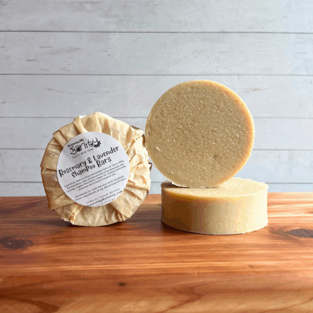 Alternate view display photo showing three round Rosemary and Lavender Shampoo bars of soap. The left bar is wrapped in an unbleached coffee filter with a label on it, the other two are unpackaged/unlabeled to show the side and face of the soap. All are sitting on a cedar board with white panel background.