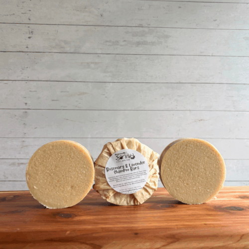 Display photo showing three round Rosemary and Lavender Shampoo bars of soap. The middle bar is wrapped in an unbleached coffee filter with a label on it, the other two are unpackaged/unlabeled to show the soap. All are sitting on a cedar board with white panel background.