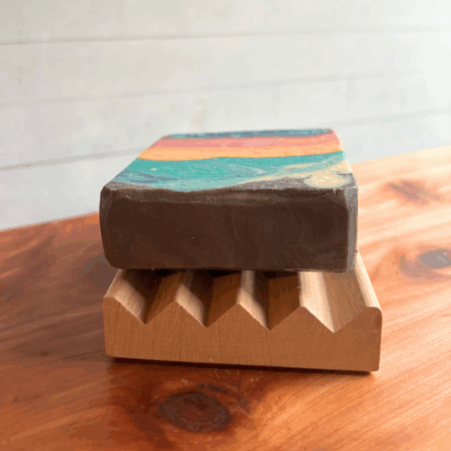 Close up display photo showing an Alder Boardwalk soap dish with a Candy Sunset bar of soap on it. Sitting on a cedar board with blurred white panel background.
