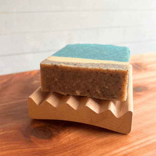 Close up display photo showing an Alder elevation soap dish with a Day at the Beach bar of soap on it. Sitting on a cedar board with blurred white panel background.