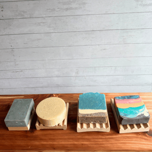 Alternate view display photo showing four sizes of natural wood soap dishes, each with a bar of soap on it. From left to right, mini Alder soap dish, square soap dish, Alder elevation soap dish, and Alder Boardwalk soap dish.
