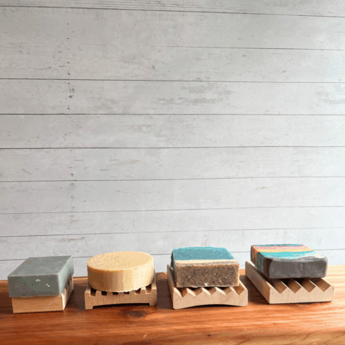 Display photo showing four sizes of natural wood soap dishes, each with a bar of soap on it. From left to right, mini Alder soap dish, square soap dish, Alder elevation soap dish, and Alder Boardwalk soap dish.