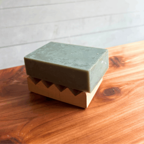 Display photo showing a mini Alder natural wood soap dish with a bar of Gardener's Soap on it, displayed on a cedar board with a white panel background.