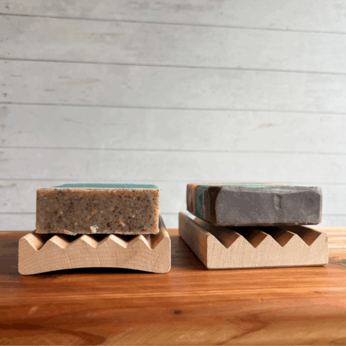 Display photo showing an Alder Elevation soap dish with a bar of soap on it, next to an Alder Boardwalk soap dish with a bar of soap on it. They are displayed next to each other to show the difference, and are displayed on a cedar board with a white panel background.