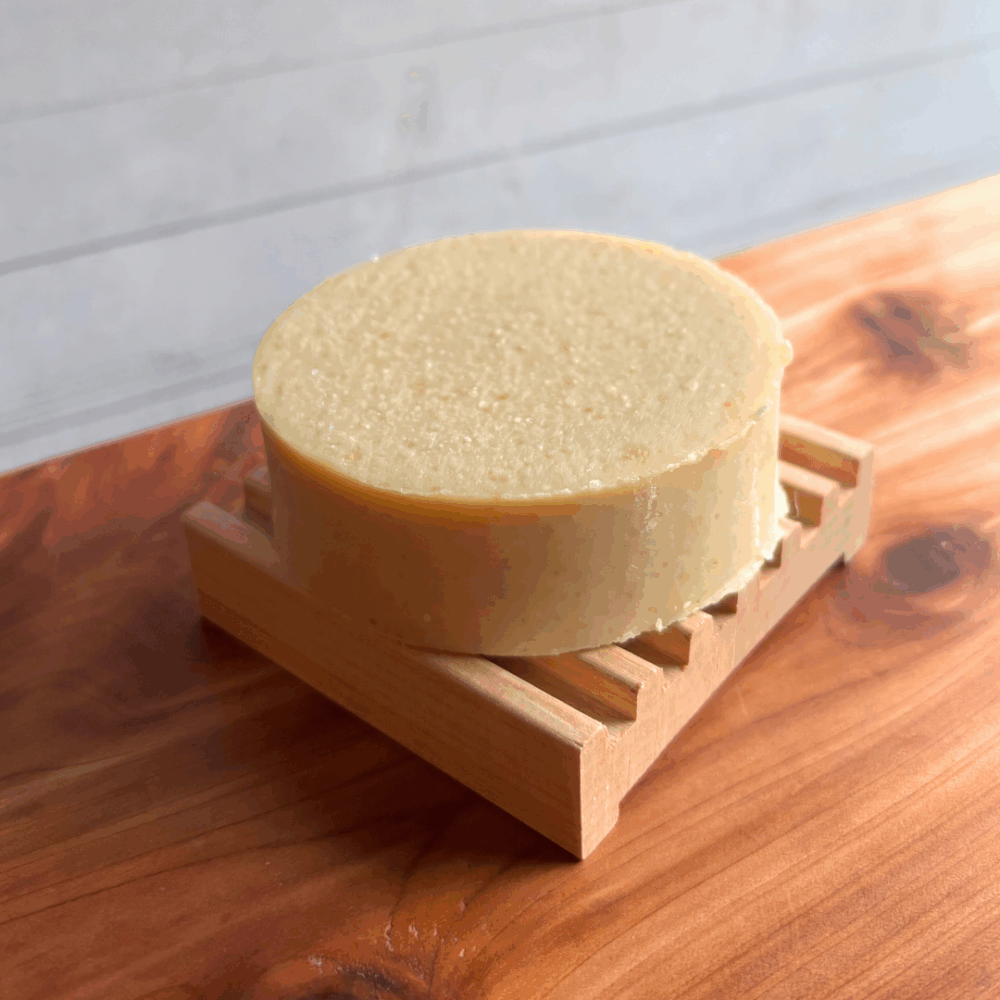 Display photo showing a shampoo bar sitting on a square natural wood soap dish, displayed on a cedar board with a white panel background.