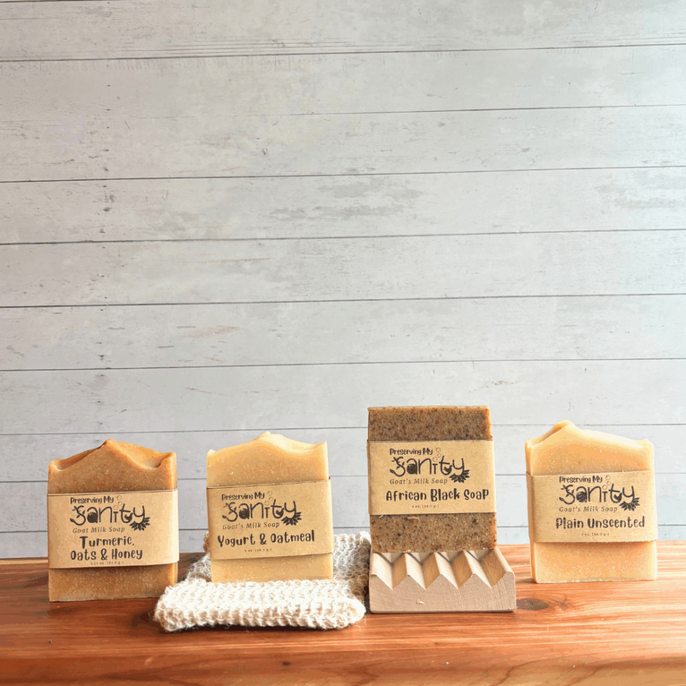 Alternative view display photo showing the contents of the Unscented Soap Sampler - half size bars of Yogurt & Oatmeal soap, African Black soap, Turmeric, Oats & Honey Soap, and Plain Unscented Soap. The sampler also comes with a natural wood soap dish and sisal soap saver bag. All of these items are displayed on a cedar board with a white panel background.
