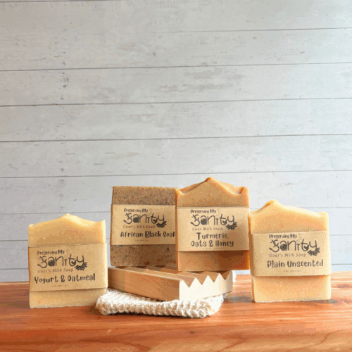 Display photo showing the contents of the Unscented Soap Sampler - half size bars of Yogurt & Oatmeal soap, African Black soap, Turmeric, Oats & Honey Soap, and Plain Unscented Soap. The sampler also comes with a natural wood soap dish and sisal soap saver bag. All of these items are displayed on a cedar board with a white panel background.
