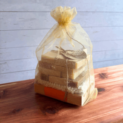 Unscented soap sampler in a gold organza bag, containing four half size bars of unscented goat milk soap varieties, a natural wood soap dish, and a sisal soap saver bag. Pictured sitting on a cedar board with white panel background.