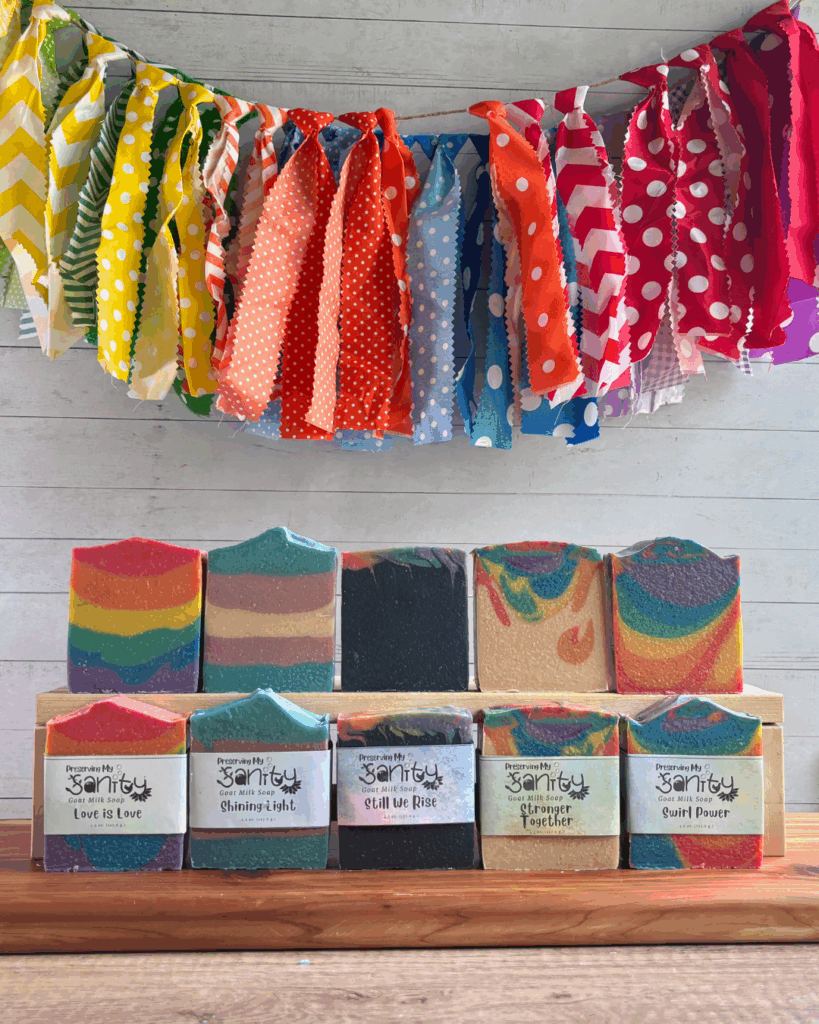 Display collage photo showing the full Pride Soap collection made by Preserving My Sanity to celebrate Pride Month 2025 and beyond. There is a colorful rainbow fabric banner in the background, and then two sets of five different kinds of Pride soaps - two each of five kinds that show varying designs, colors, and labels.