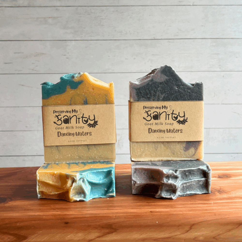 Four bars of Dancing Waters goat milk soap, made in two color variations. One bar is blue, gold, and uncolored while the other is purple, charcoal, and uncolored. Bars are displayed to show the tops and the fronts with soap labels, situated on a cedar bath board with a white paneled background.