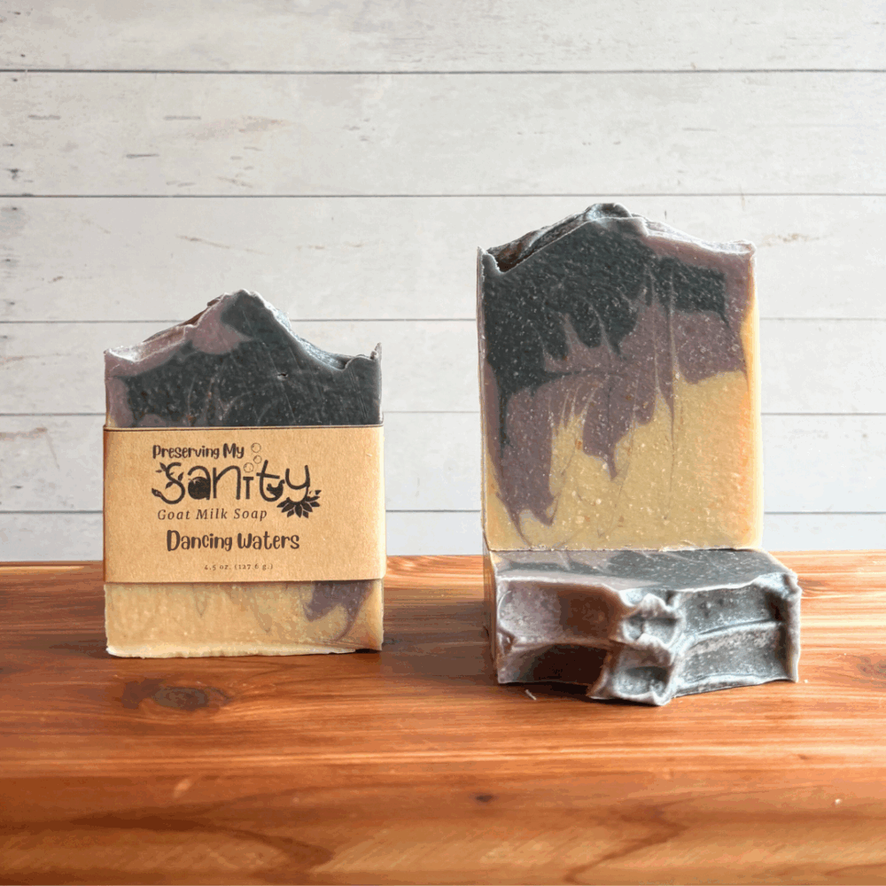 Three bars of Dancing Waters goat milk soap. Soap is a unique swirl of purple, charcoal, and uncolored soap. Two bars are displayed to show the design and top of the bars, while the third shows the label.
