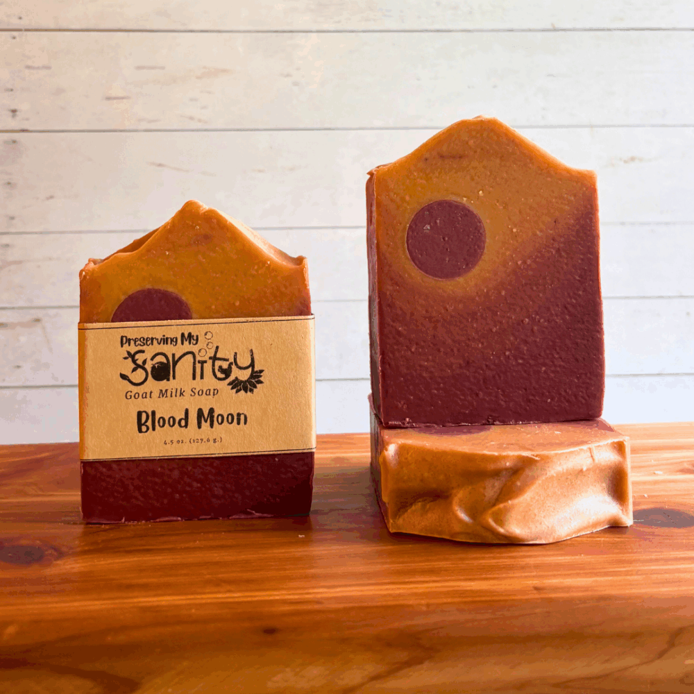 Three bars of Blood Moon goat milk soap. The soap was inspired by a photo and created with an ombre pour from dark red to lighter red to orange to gold. Two bars are displayed to show the design and top of the bars, while the third shows the label.