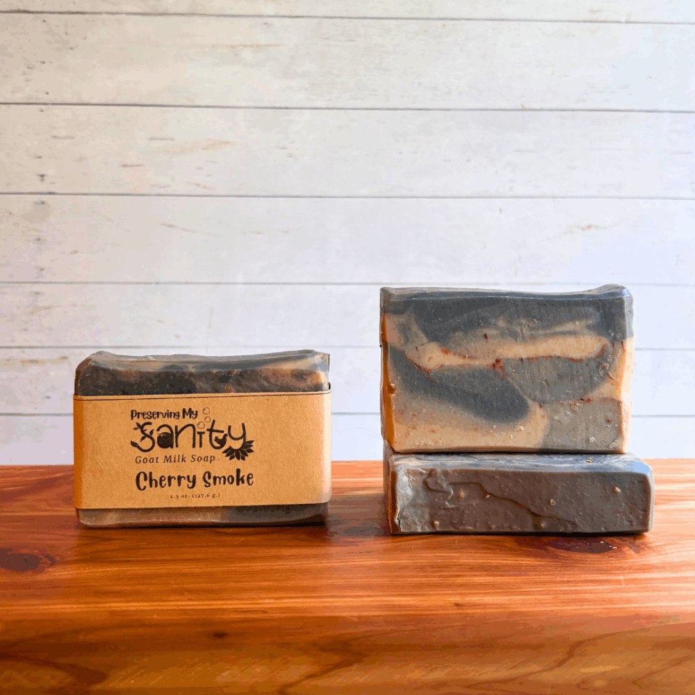 Three bars of Cherry Smoke goat milk soap. The soap was inspired by a photo and created with a marbled pattern of gray, pink, and white. Two bars are displayed to show the design and top of the bars, while the third shows the label.