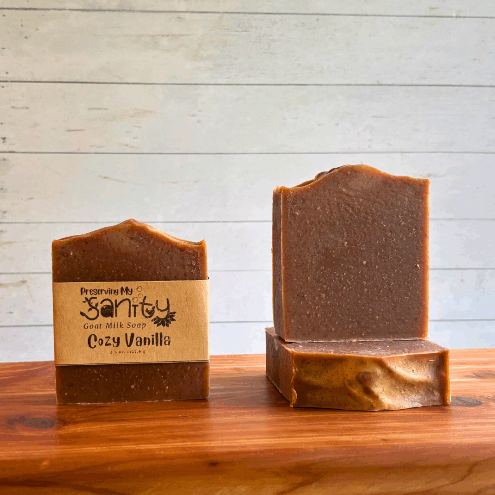 Three bars of Cozy Vanilla goat milk soap. The soap is a dark camel brown color. Two bars are displayed to show the design and top of the bars, while the third shows the label.