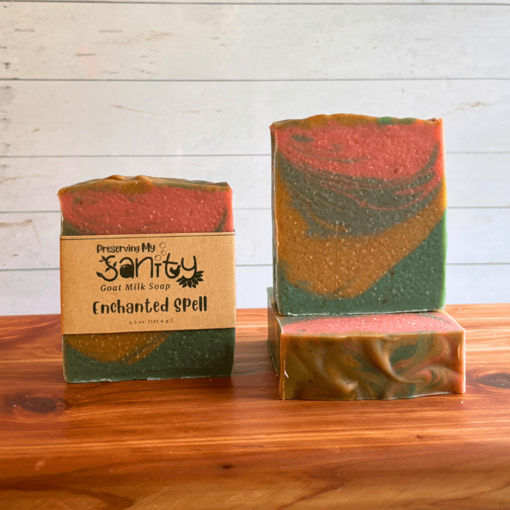 Three bars of Enchanted Spell goat milk soap displayed in a row on a cedar bath board with a white paneled background. Two bars are displayed to show the swirly layers of forest green, gold, sea blue, and coral found in this unique paranormal themed goat milk soap. The third bar is laid flat on the board to display the textured and swirled soap top.