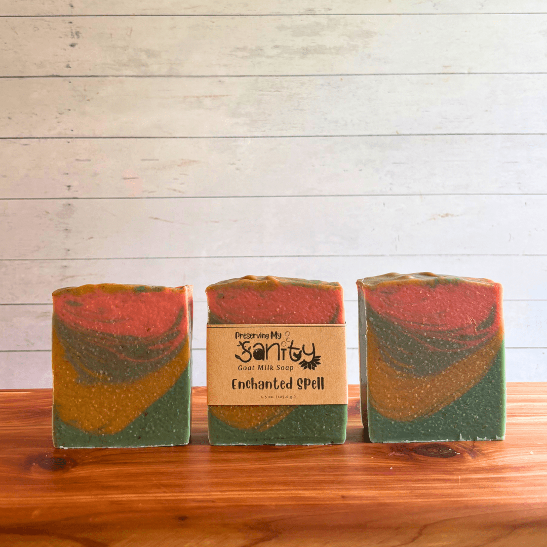 Three bars of Enchanted Spell goat milk soap. This soap was poured in a one pot wonder style and features swirly layers of forest green, gold, sea blue, and coral. Two bars are displayed to show the design and top of the bars, while the third shows the label.