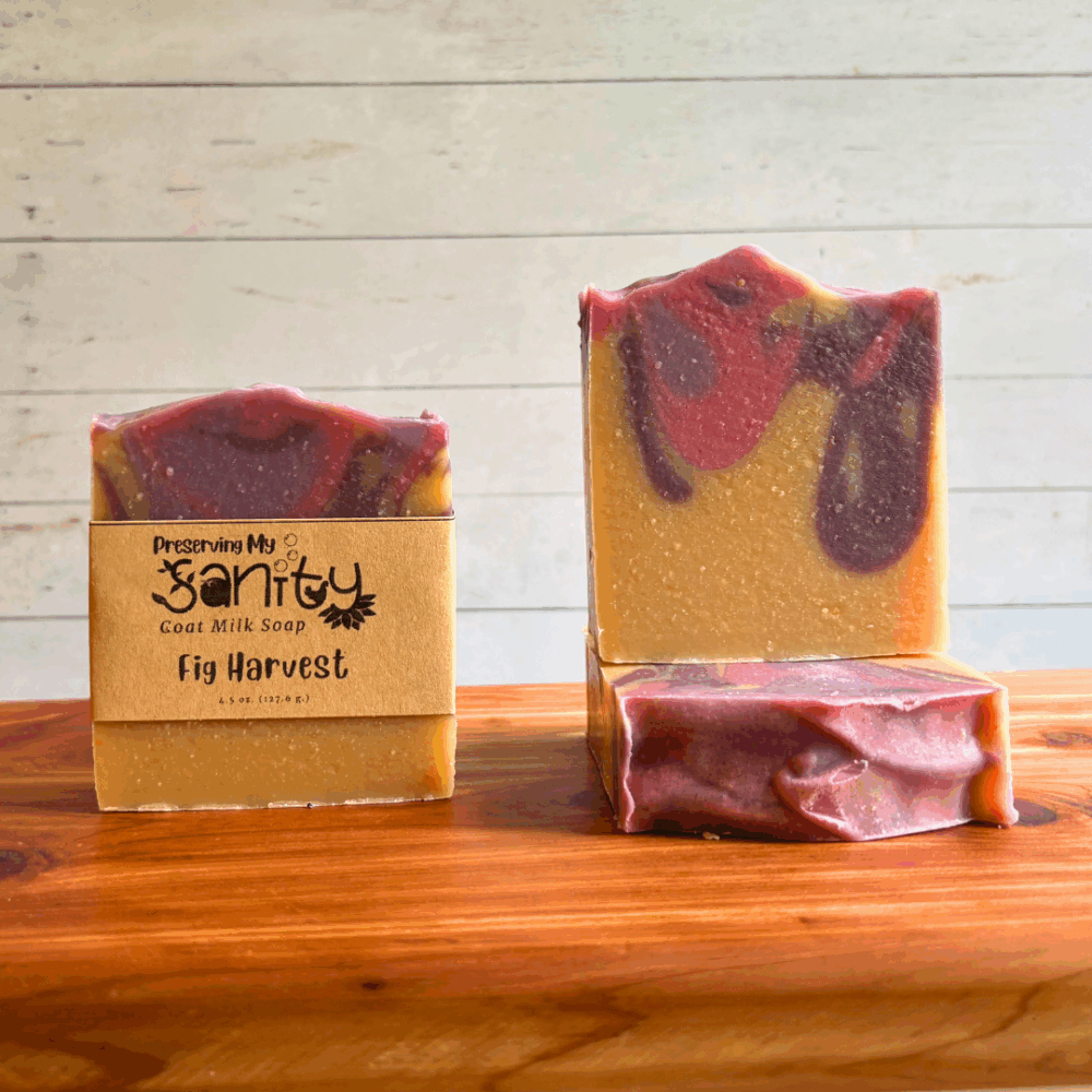 Three bars of Fig Harvest goat milk soap. This soap has an uncolored base, with purple, pink, and lavender drop swirled into it. Two bars are displayed to show the design and top of the bars, while the third shows the label.