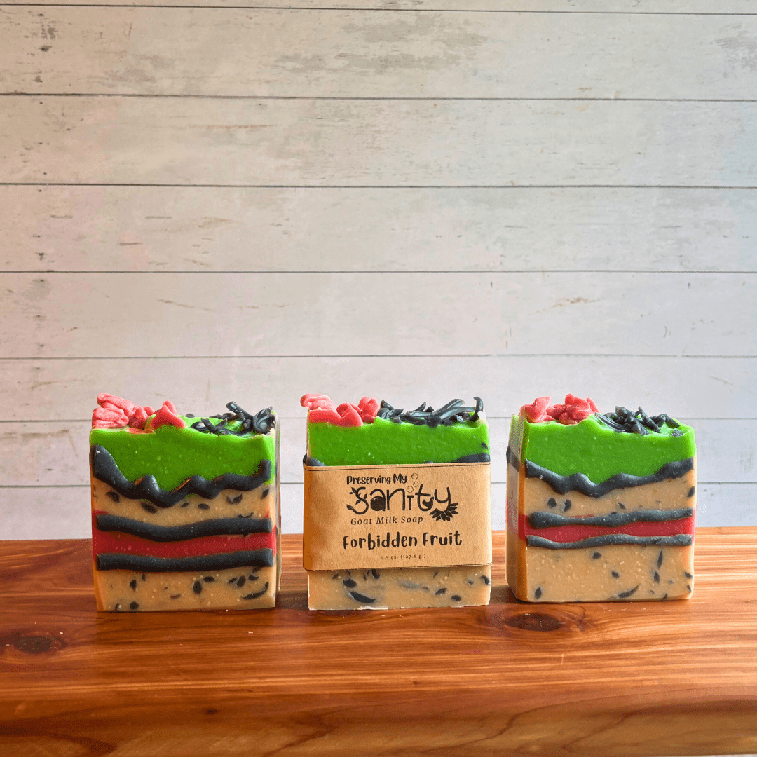 Three bars of Forbidden Fruit goat milk soap displayed in a row on a cedar bath board with a white paneled background. Two bars are displayed to show the fun design of this comic soap, with layers of white with charcoal sprinkles, red frosting, lime green frosting, funky piping on top, and black piping on the front. It looks spooky AND delicious! The third bar of soap has a label on it.