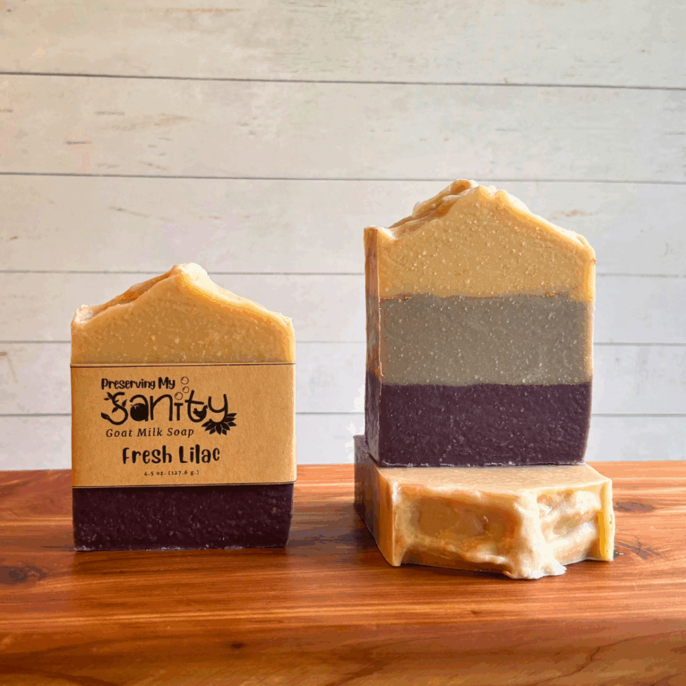 Three bars of Fresh Lilac goat milk soap. This soap was carefully poured in layers of dark purple, lavender, and white - with gold pencil lines in between. Two bars are displayed to show the design and top of the bars, while the third shows the label.