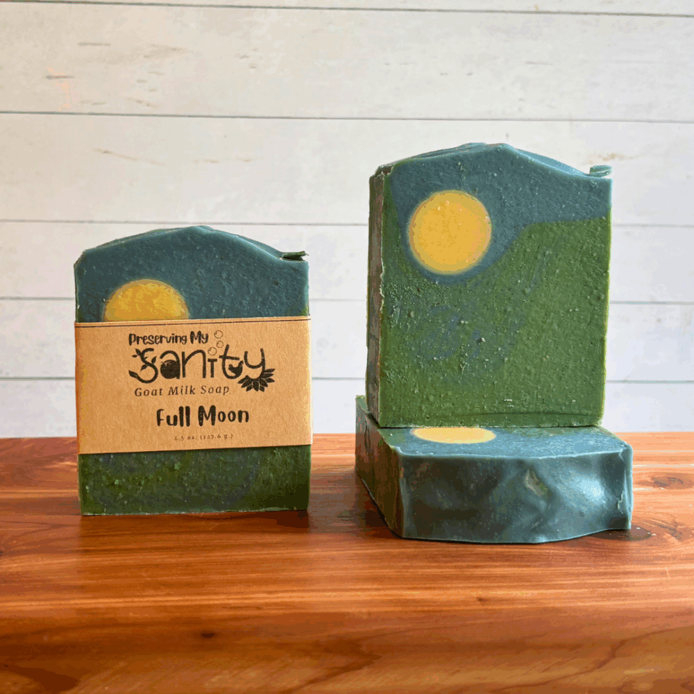 Three bars of Full Moon goat milk soap. This soap was carefully poured to look like a hillside with a dark night sky with a full moon, and the fragrance is mahogany teakwood. Two bars are displayed to show the design and top of the bars, while the third shows the label.