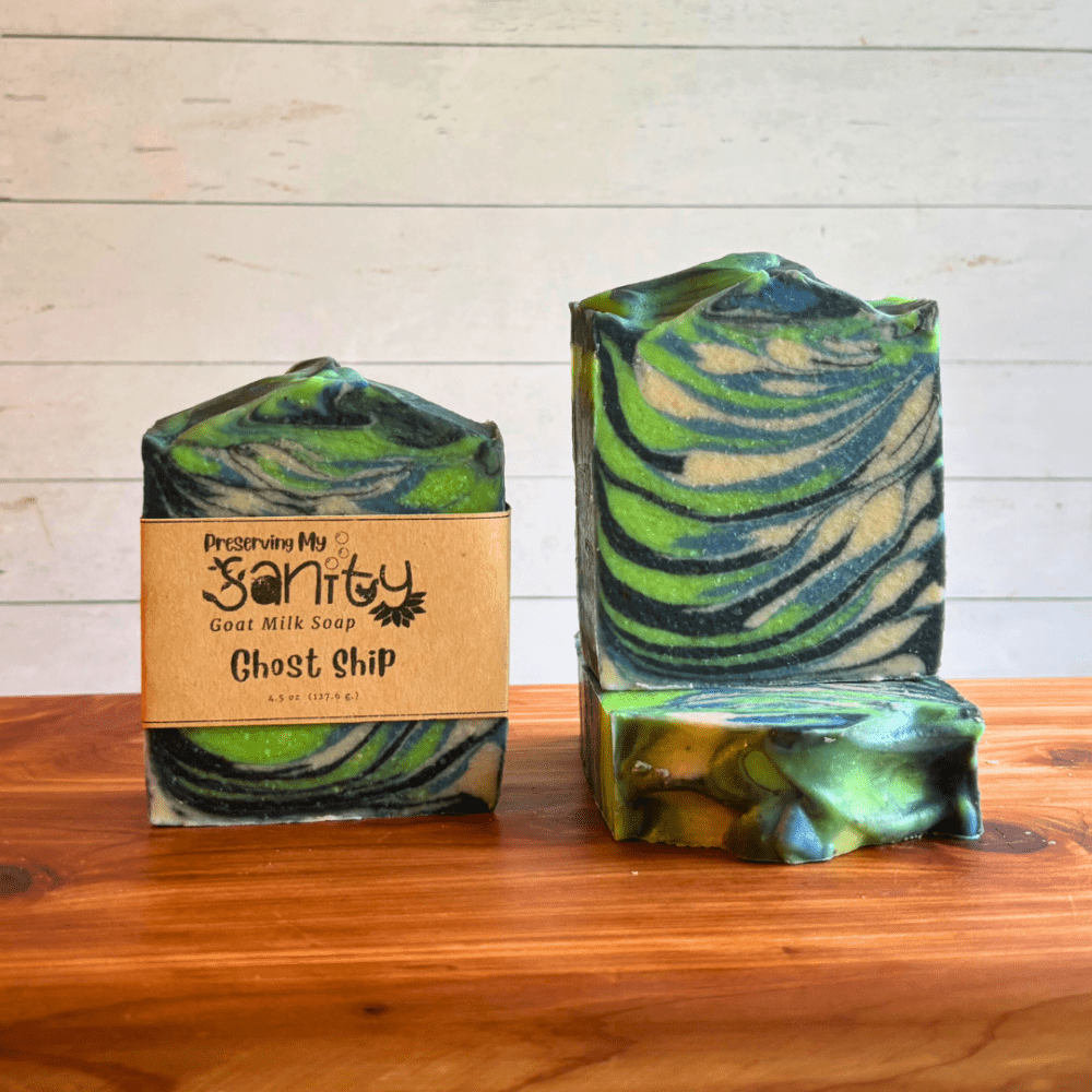 Three bars of Ghost Ship goat milk soap displayed on a cedar bath board with a white paneled background. Two bars are displayed to show the wispy swirls of black, blue, lime green, and white in this paranormal-themed soap. The third bar is laying flat on the board to display the textured soap top.