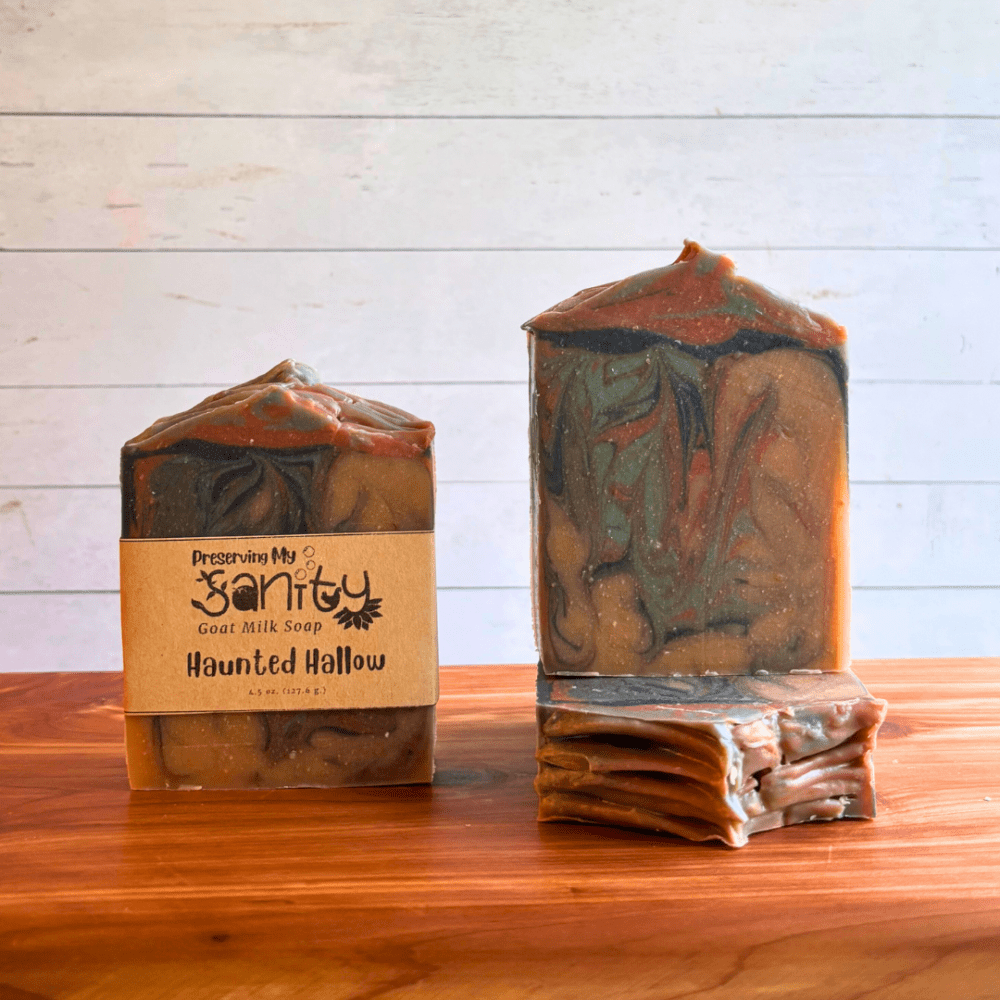 Three bars of Haunted Hallow goat milk soap. This soap has an eerie blend of light brown, dark brown, green, and black swirls that are perfect for the spooky season. Two bars are displayed to show the design and top of the bars, while the third shows the label.
