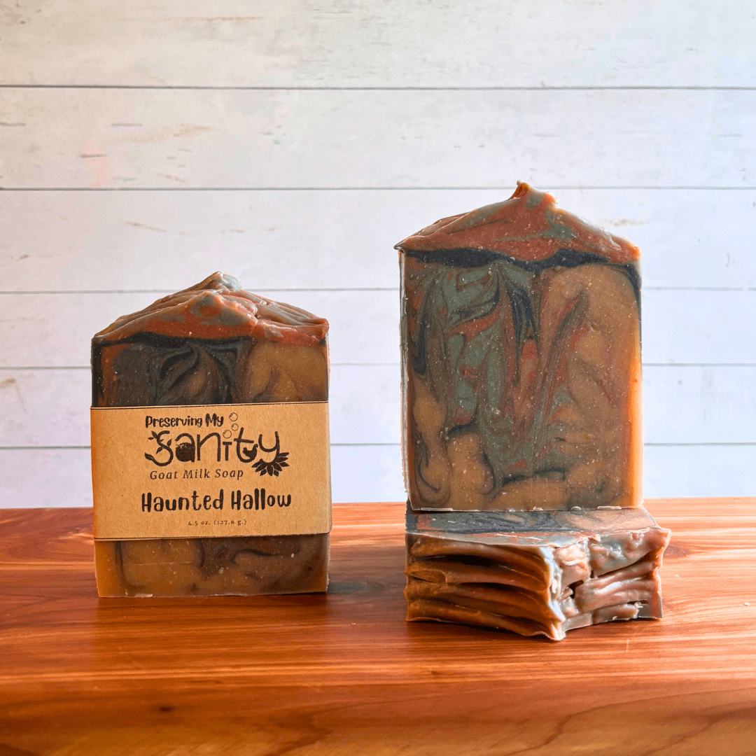 Three bars of Haunted Hallow goat milk soap. This soap has an eerie blend of light brown, dark brown, green, and black swirls that are perfect for the spooky season. Two bars are displayed to show the design and top of the bars, while the third shows the label.