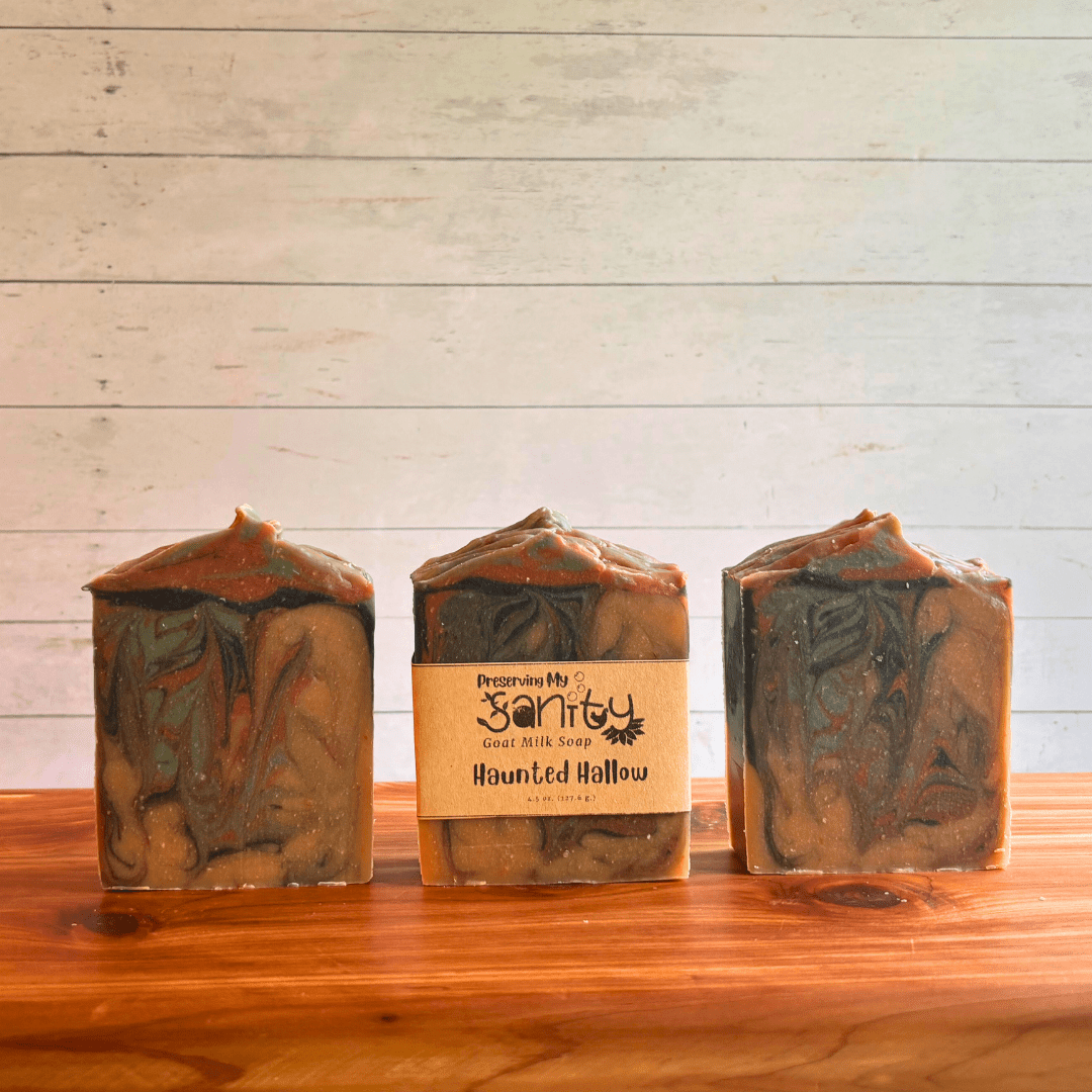 Three bars of Haunted Hallow goat milk soap displayed in a row on a cedar bath board with a white paneled background. Two bars are displayed to show the spooky swirls of brown, beige, green, and charcoal in this paranormal-themed soap. The third bar of soap has a label on it.