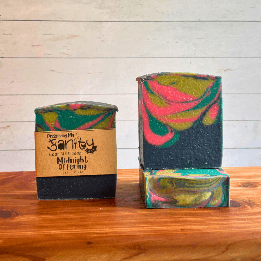 Three bars of Midnight Offering goat milk soap. This soap has a dark blue base and contains drop swirls of hot pink, lime green, and turquoise blue. Two bars are displayed to show the design and top of the bars, while the third shows the label.