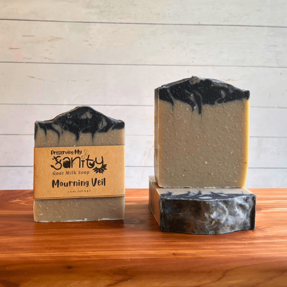 Three bars of Mourning Veil goat milk soap. This soap has a silver and lavender base and is topped with a swirly black layer. Two bars are displayed to show the design and top of the bars, while the third shows the label.