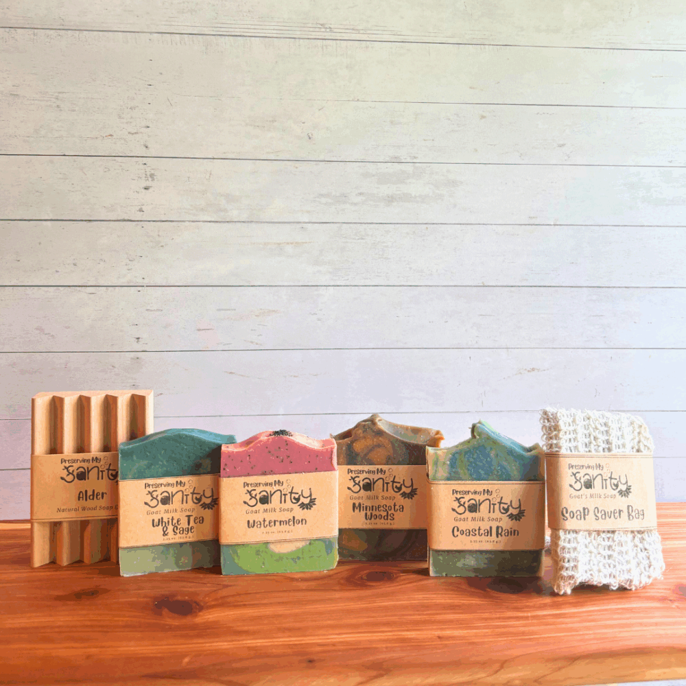 Display photo of the items included in a Bestsellers Soap Sampler - four half size bars of destination-themed soap, a sisal fiber soap saver bag, and a natural wood soap dish. All are pictured upright on a cedar bath board, with a white paneled background.