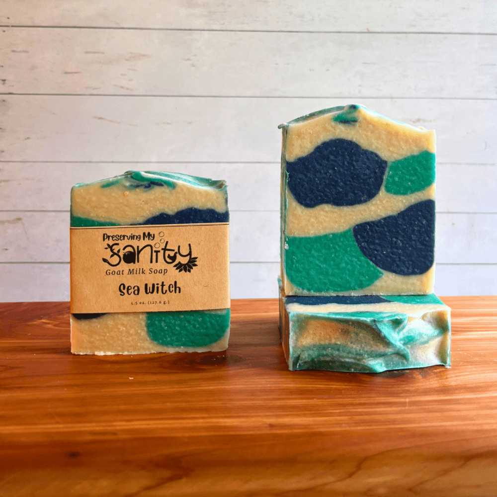 Three bars of Sea Witch goat milk soap. The soap design was inspired by a skinny shimmy, but I used a regular mold and fewer colors so it turned out in these fun shapes of aqua blue, navy blue, and white. Two bars are displayed to show the design and top of the bars, while the third shows the label.