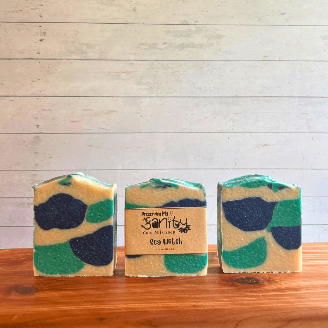 Three bars of Sea Witch goat milk soap displayed in a row on a cedar bath board with a white paneled background. Two bars are displayed to show the funky shapes of aqua blue, navy blue, and white found in this unique paranormal themed goat milk soap. The third bar of soap has a label on it.