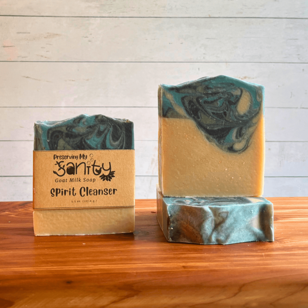 Three bars of Spirit Cleanser goat milk soap. The bottom half of the soap bars is uncolored, while the top is a unique swirled pattern of gray, aqua blue, and black. Two bars are displayed to show the design and top of the bars, while the third shows the label.