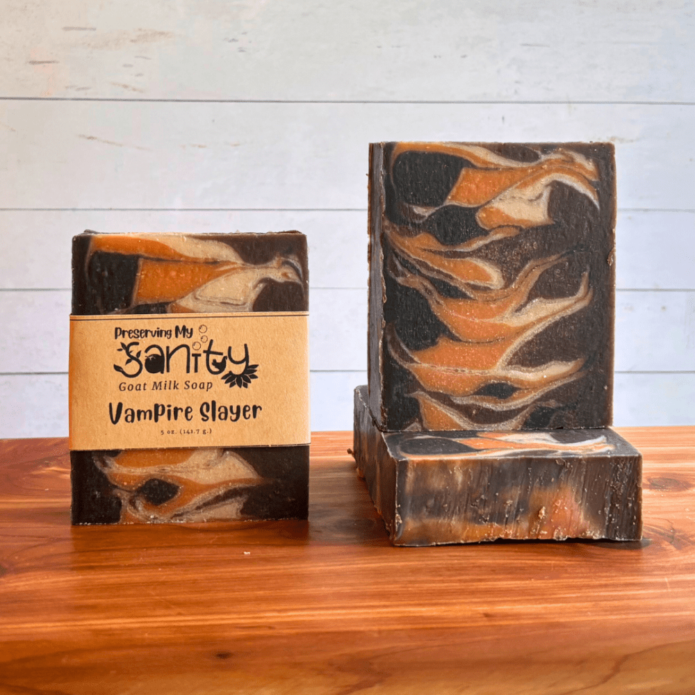 Three bars of Vampire Slayer goat milk soap. The soap is a Taiwan swirl design of black, brown, orange, and white. Two bars are displayed to show the design and top of the bars, while the third is laid flat on the board to display the soap top.